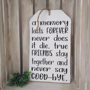 May include: White wooden sign with a tag shape, featuring the quote "a memory lasts FOREVER never does it die. true FRIENDS stay together and never say GOOD-BYE." The sign has a jute rope for hanging and a small green plant.