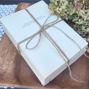 May include: A stack of aged book pages tied with twine, creating a rustic aesthetic. The pages are off-white, with visible text. The twine is tied in a bow. The stack rests on a wooden tray, with dried flowers and beads in the background.