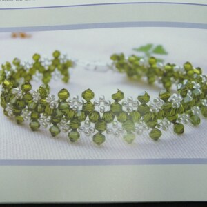May include: A beaded bracelet featuring olive green and clear beads. The bracelet has a delicate floral design with a silver clasp. The beads are arranged in a repeating pattern, creating a textured look. The bracelet is displayed on a white surface.