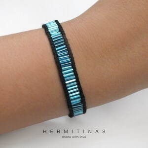 May include: A close-up of a beaded bracelet. The bracelet features rows of small, rectangular, turquoise beads set against a black background. The bracelet is worn on a wrist. The brand name "HERMITINAS" and the phrase "made with love" are at the bottom.