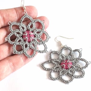 May include: A pair of handmade silver-grey flower-shaped earrings with a pink centre. The earrings are made of a delicate, woven material and feature a hook closure. The floral design is intricate and detailed, with multiple petals.