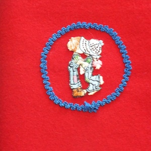 May include: A red felt fabric with a blue embroidered circle. Inside the circle is a colorful embroidered image of two children kissing. The children are wearing overalls and hats. The embroidery is detailed and the colors are vibrant.