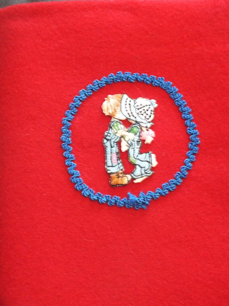 May include: A red felt fabric with a blue embroidered circle. Inside the circle is a colorful embroidered image of two children kissing. The children are wearing overalls and hats. The embroidery is detailed and the colors are vibrant.