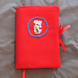 May include: A bright red book cover with a blue embroidered circle featuring a small figure. A red ribbon is attached to the cover. The cover is made of a soft, felt-like material.
