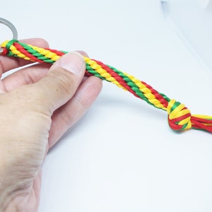 Könnte beinhalten: Ein geflochtener Schlüsselanhänger mit einem Metallring. Der Schlüsselanhänger besteht aus roten, gelben und grünen Kordeln, die am Ende mit passenden Quasten verknotet sind. Das Design wird von einer Hand gehalten.