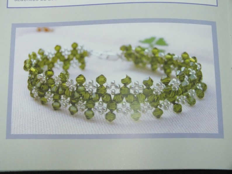 May include: A beaded bracelet featuring olive green and clear beads. The bracelet has a delicate floral design with a silver clasp. The beads are arranged in a repeating pattern, creating a textured look. The bracelet is displayed on a white surface.