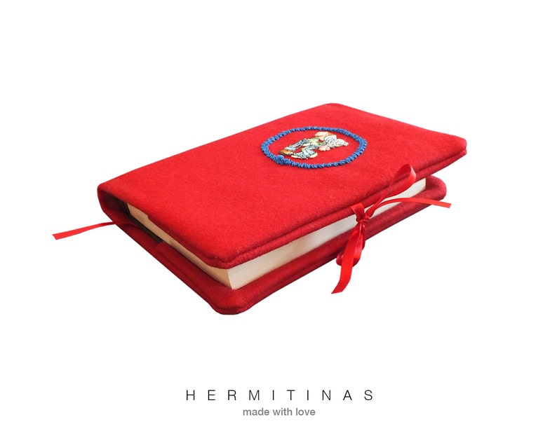 May include: A red book cover with an embroidered design in a blue circle. The cover is tied with a red ribbon and has a red bookmark. The text "HERMITINAS made with love" is at the bottom.