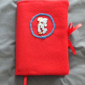 May include: A bright red book cover with a blue embroidered circle featuring a couple. A red ribbon bookmark and tie are attached. The cover is made of a soft, felt-like material.