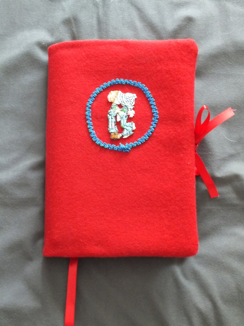 May include: A bright red book cover with a blue embroidered circle featuring a couple. A red ribbon bookmark and tie are attached. The cover is made of a soft, felt-like material.