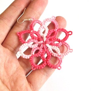 以下が含まれることがあります： ピンクと白の色合いの手作りの花型のイヤリング。イヤリングは、銀色のフックが付いた繊細なオープンワークデザインが特徴です。複雑なパターンは、ユニークでフェミニンなアクセサリーを作成します。