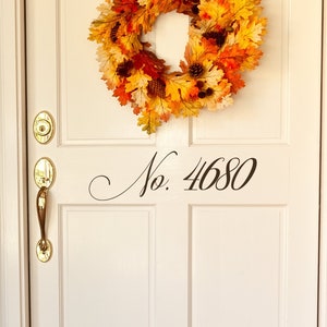 May include: A white front door with a fall-themed wreath made of orange, yellow, and brown leaves. The door has a brass door handle and a brass door knocker. The house number "No. 4680" is written in black cursive on the door.