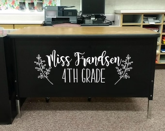 May include: A black rectangular teacher desk with a light brown top and silver legs. The front panel features white vinyl lettering that reads Miss Frandsen 4TH GRADE, flanked by two delicate white line-art botanical branch illustrations.