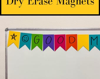 May include: A set of colorful dry erase magnets shaped like pennant flags, arranged in a row on a white board. The magnets are yellow, teal, green, purple, pink, orange, and blue, featuring hand-drawn black ink symbols and letters spelling GOOD M.