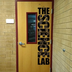 May include: A light wood door with a dark red frame and a rectangular window. A black vinyl decal on the door features the text THE SCIENCE LAB, with the word SCIENCE filled with various small icons including a whale, skull, atom, and plant.