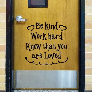 May include: A wooden door featuring a black vinyl decal with the text "Be kind Work hard Know that you are Loved" flanked by two small heart icons and a decorative swirly line underneath, all centered on the door surface below a silver handle.