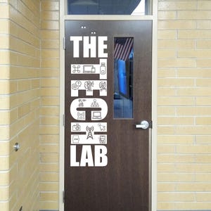 May include: A brown door with a rectangular window, featuring white text and icons that read "THE TECH LAB." The door has a silver handle and is set in a yellow brick wall. An American flag is visible through the window.