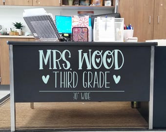 May include: A black desk front panel featuring light blue vinyl lettering that reads MRS. WOOD THIRD GRADE with heart icons and a horizontal line. Below the text, the measurement 30" WIDE is printed in a thin, white, handwritten-style font.