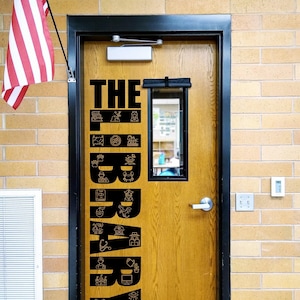 May include: A wooden door with a black vertical decal that reads THE LIBRARY, featuring various small line-art icons integrated into the letters. The door has a rectangular window, a silver handle, and a black frame, with an American flag nearby.