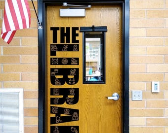 May include: A wooden door with a black vertical decal that reads THE LIBRARY, featuring various small line-art icons integrated into the letters. The door has a rectangular window, a silver handle, and a black frame, with an American flag nearby.