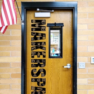 May include: A wooden door with a black vertical decal reading MAKERSPACE, featuring various icons like a lightbulb, gears, and math symbols integrated into the letters. The door has a silver handle, a rectangular window, and a US flag to the left.