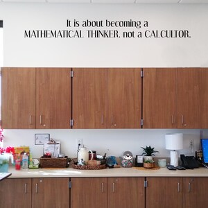 May include: A wall decal with black lettering that reads: It is about becoming a MATHEMATICAL THINKER, not a CALCULTOR. The text is positioned above a set of brown wooden cabinets with silver handles, which are mounted on a white wall.