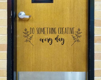 May include: A wooden door featuring a black vinyl decal that reads DO SOMETHING CREATIVE every day, flanked by two botanical leaf illustrations. The door includes a silver metal handle and a rectangular window at the top with a metal kick plate.
