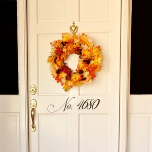 May include: A white front door featuring a brass door knocker, a brass handle, and black house numbers reading No. 4680. A circular autumn wreath made of orange, yellow, and red maple leaves with small brown pinecones hangs from the door knocker.