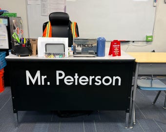 May include: A black teacher's desk with the name Mr. Peterson in large white letters on the front. The desk surface holds a pencil holder, a white organizer, a silver metal box labeled PENCILS, a blue canister, and a red sign with white text.