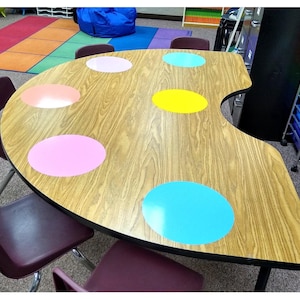May include: A kidney-shaped table with a light brown wood grain laminate surface featuring six solid-colored circular decals in pink, light pink, yellow, light blue, and teal. The table is surrounded by several dark maroon plastic chairs.