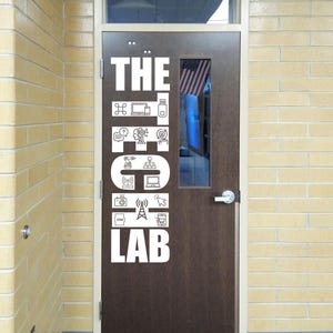May include: A brown door with white text and icons that read "THE TECH LAB." The door has a small window with a glimpse of an American flag. The door is set in a yellow brick wall.