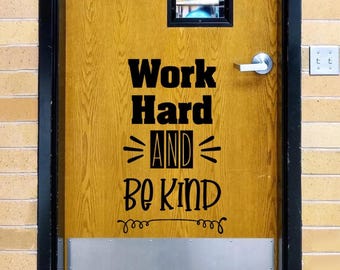 May include: A wooden door featuring a black vinyl decal with the text Work Hard AND BE KIND. The door has a silver metal handle, a small rectangular window at the top, and a silver kick plate at the bottom, set within a black frame.