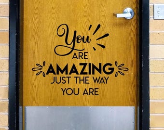 May include: A wooden door featuring a black vinyl decal with the text "You ARE AMAZING JUST THE WAY YOU ARE" in a mix of script and sans-serif fonts. The door includes a silver metal handle and a silver kick plate at the bottom of the door panel.