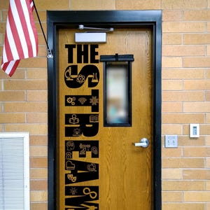 May include: A wooden door with a black frame featuring a vertical black decal that reads THE STREAM LAB, decorated with various icons representing science, technology, engineering, art, and mathematics, positioned next to an American flag.