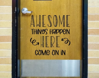 May include: A wooden door featuring a black vinyl decal with the text "AWESOME THINGS HAPPEN HERE COME ON IN" in a mix of stylized fonts. The door includes a silver metal handle and a silver kick plate at the bottom, set within a dark frame.