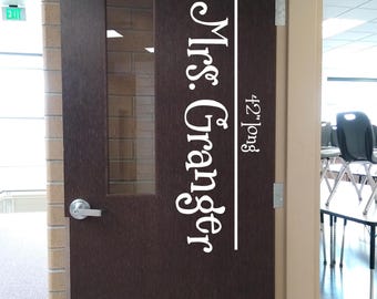 May include: A dark brown wooden door featuring a vertical white vinyl decal that reads Mrs. Granger and 42" long. The door includes a rectangular glass window, a silver metal handle, a top-mounted door closer, and a silver kick plate at the bottom.