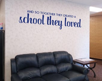 May include: A wall decal featuring the dark blue text "AND SO TOGETHER THEY CREATED A school they loved" in a mix of block and script fonts. The lettering is applied to a light-colored wall above a black leather three-cushion sofa.