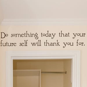 May include: A black wall decal featuring the quote "Do something today that your future self will thank you for." in a stylized, rounded serif font. The lettering is applied directly to a light cream-colored wall above a white door frame.