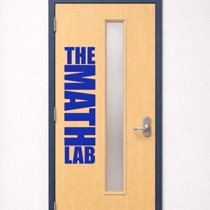 May include: A light wood-toned door with a dark blue frame and a vertical rectangular glass window. The door features bold blue text applied to the surface that reads THE MATH LAB, stacked vertically, and includes a silver metal handle and kick plate.