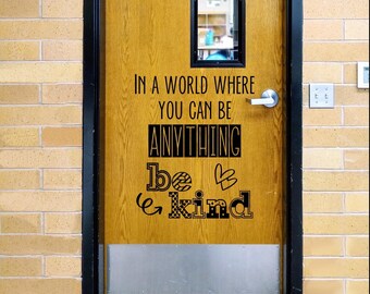 May include: A wooden door featuring a black vinyl decal with the text: IN A WORLD WHERE YOU CAN BE ANYTHING, BE KIND. The word ANYTHING is in a bold block font, while the words be kind feature decorative patterns, including hearts and swirls.