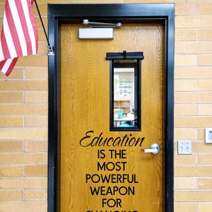 May include: A wooden door with a black frame and a rectangular window, featuring the black text: Education IS THE MOST POWERFUL WEAPON FOR CHANGING THE WORLD. A silver door handle and a metal kick plate are visible, with an American flag to the left.