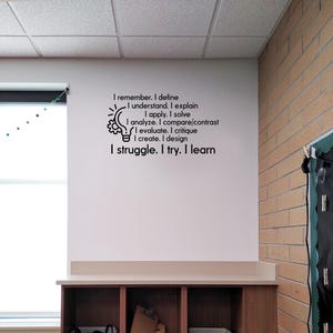 May include: A black wall decal featuring a lightbulb icon with a gear inside, accompanied by the text: I remember. I define. I understand. I explain. I apply. I solve. I analyze. I compare/contrast. I evaluate. I critique. I create. I design. I struggle. I try. I learn.