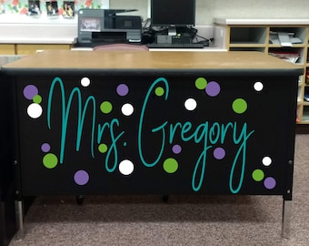 May include: A black desk with a brown top and silver legs, featuring the name Mrs. Gregory in large teal cursive script. The front panel is decorated with a pattern of scattered white, lime green, and purple circular polka dots of varying sizes.