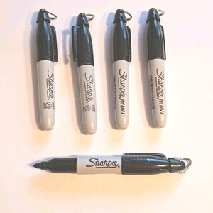 May include: A collection of five black and silver Sharpie Mini permanent markers. Four markers are upright, and one is horizontal. Each marker has a silver keyring attached to the cap and the brand name "Sharpie" printed on the barrel.