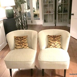 May include: Two armless chairs upholstered in a cream-coloured fabric with a subtle chevron pattern, each featuring a square throw pillow with a brown and black tiger stripe print and dark wooden legs, positioned side by side in a room.
