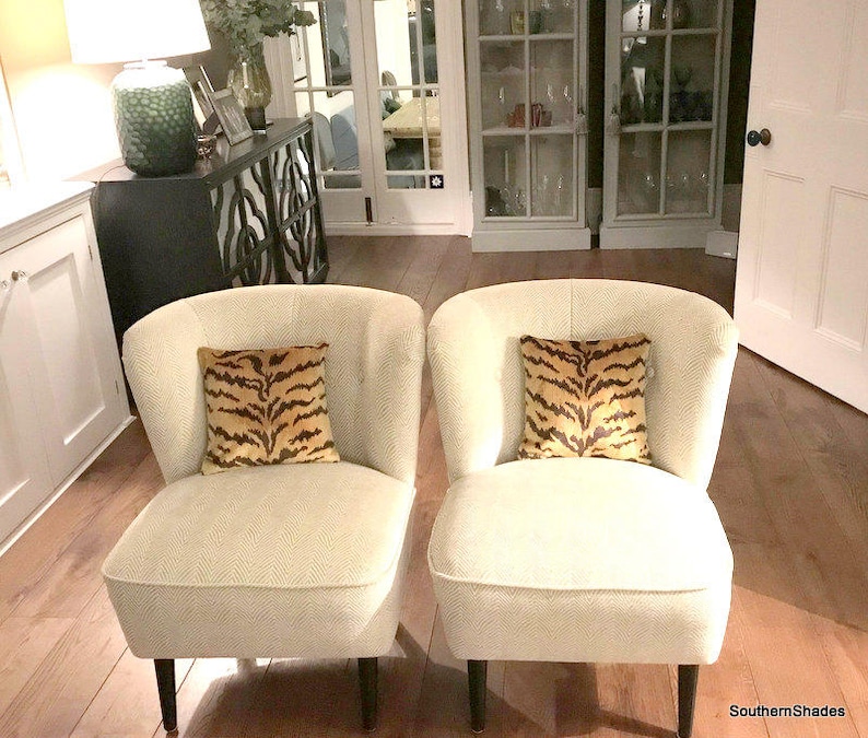 May include: Two armless chairs upholstered in a cream-coloured fabric with a subtle chevron pattern, each featuring a square throw pillow with a brown and black tiger stripe print and dark wooden legs, positioned side by side in a room.