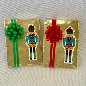 Puede incluir: Dos cajas de regalo doradas con lazos de cinta roja y verde. Cada caja tiene una pegatina de cascanueces con un sombrero rojo y verde y un tambor.