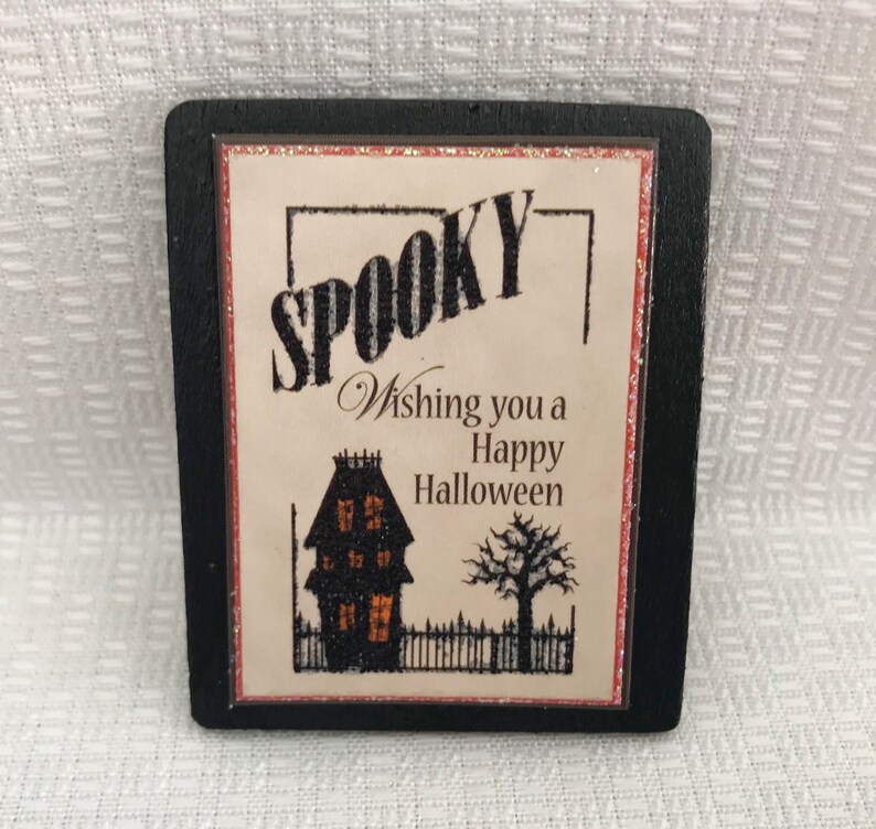 Pode incluir: Um sinal enquadrado em preto com fundo branco e guarni&ccedil;&atilde;o de glitter vermelho. O sinal diz "Spooky" em letras pretas e brancas listradas. Abaixo da palavra "Spooky" est&aacute; o texto "Wishing you a Happy Halloween". Uma silhueta preta de uma casa com janelas laranja e uma silhueta preta de uma &aacute;rvore est&atilde;o abaixo do texto.