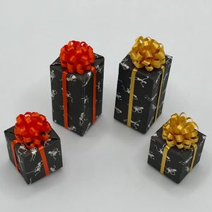 May include: Four small gift boxes wrapped in black paper with a skeleton print. Each box is adorned with a colorful bow. Two boxes have orange bows and orange ribbon, while the other two have gold bows and gold ribbon.