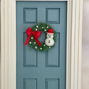 May include: A miniature blue door with a white frame, adorned with a Christmas wreath. The wreath features a red bow, white accents, and a small snowman wearing a red hat. A festive holiday decoration.