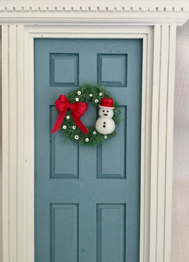 May include: A miniature blue door with a white frame, adorned with a Christmas wreath. The wreath features a red bow, white accents, and a small snowman wearing a red hat. A festive holiday decoration.